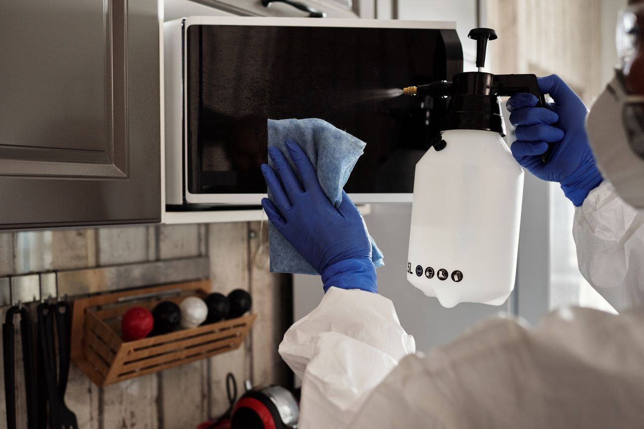 heros-img A person wearing protective equipment is cleaning a microwave indoors with a spray bottle and cloth.
